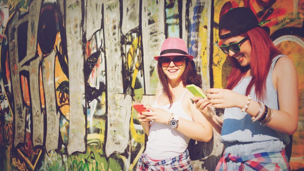 two girls leaning on graffiti looking at phones